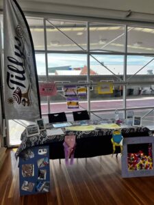 A booth with a "Tilley" banner features a table decorated with crafts, photos, and colorful signs. Large windows reveal a view of buildings outside.