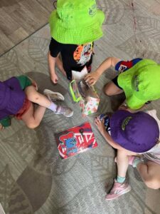 Four children wearing brightly colored hats sit on a patterned floor, engaged in opening a colorful gift. The scene exudes curiosity and excitement.