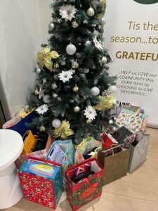 A decorated Christmas tree with gold and white ornaments has numerous colorful gift bags beneath it. Beside the tree, a sign encourages gratitude and community giving, creating a festive and charitable atmosphere.