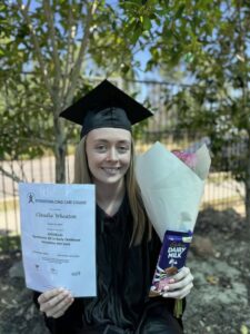 Young woman in graduation cap and gown holds a certificate, bouquet, and chocolate bar, smiling outdoors with trees in the background, conveying joy.