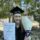 Young woman in graduation cap and gown holds a certificate, bouquet, and chocolate bar, smiling outdoors with trees in the background, conveying joy.