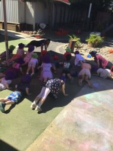 Children in colorful hats crouch on hands and knees outdoors, engaging in a playful activity. A sunny, cheerful day with scattered toys in the background.