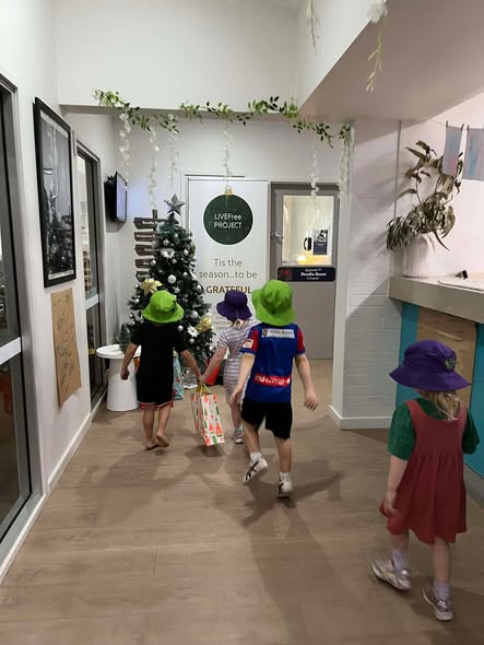 Children wearing colorful hats walk towards a decorated Christmas tree in a hallway. The atmosphere is festive and cheerful, with greenery above.
