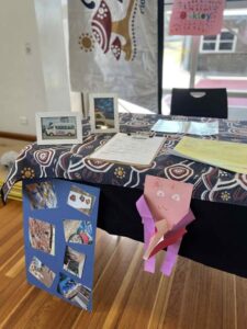 A table with a patterned cloth displays photos, paperwork, and a pink paper craft. A blue board with images is attached to the table. The setting is bright and cheerful.