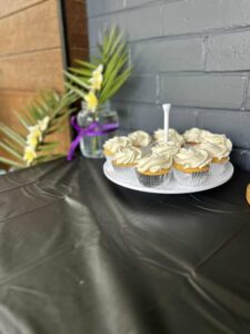 A plate of cupcakes with white frosting is on a black tablecloth. A jar with palm leaves and yellow flowers tied with a purple ribbon is in the background.