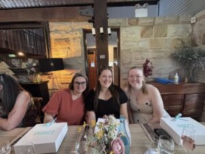 Three women sit smiling at a wooden table set with flowers and glasses in a rustic room with stone walls. Two white gift boxes are in front of them.