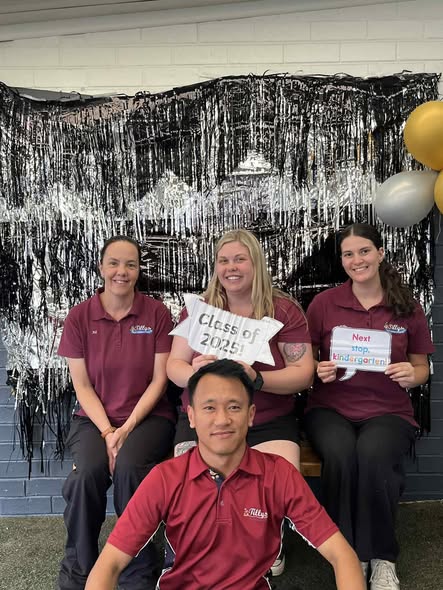 Four people in maroon shirts sit in front of a shiny black and silver backdrop. Two hold signs: “Class of 2023!” and “Next stop: Kindergarten!” Balloons float nearby.