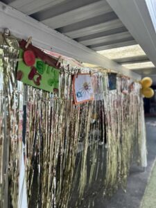 Gold tinsel hangs under a roof, with colorful child artwork, including a sun and animals, clipped above. Balloons in the background add festivity.