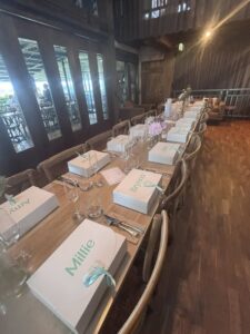 A long dining table set with white gift boxes tied with blue ribbons, each bearing a name in green. Elegant wood interior and soft ambient lighting.