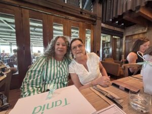 Two women seated at a wooden table in a cozy restaurant, smiling. They are surrounded by gift boxes. The atmosphere is warm and inviting.