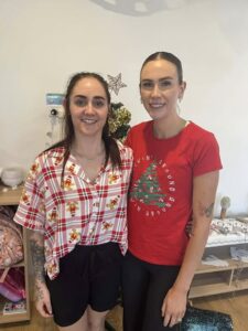 Two women smiling in a festive setting. One wears a plaid shirt with holiday motifs; the other, a red tee with a Christmas tree design. A joyful, warm atmosphere.