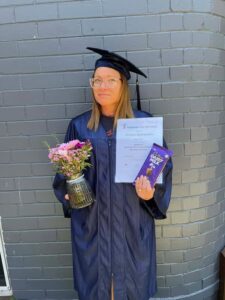 Tillys Waratah Graduate in cap and gown holds a certificate, flowers in a glass jar, and a chocolate bar, standing against a gray brick wall, smiling slightly.