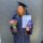 Graduate in cap and gown holds a certificate, flowers in a glass jar, and a chocolate bar, standing against a gray brick wall, smiling slightly.
