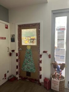 Tillys Rutherford A festive door decorated with a paper Christmas tree, red and white tinsel, and candy cane shapes. A sign says "Reindeer Parking Only." Bright holiday atmosphere.