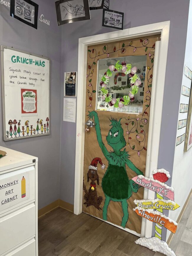Tillys Rutherford Classroom door decorated for Christmas with Grinch theme. Features a large Grinch drawing, wreath, dog in a Santa hat, and Whoville signs in festive colors.