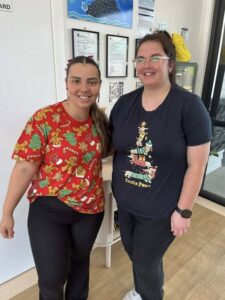 Two people smile at the camera, wearing festive holiday shirts. One has a gingerbread and tree pattern, the other a Santa climbing a tree. Cozy indoor setting.
