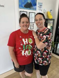 Two people smiling indoors, both wearing festive shirts; one red with "Ho Ho Ho" text, the other with dogs in Santa hats, conveying a joyful atmosphere.
