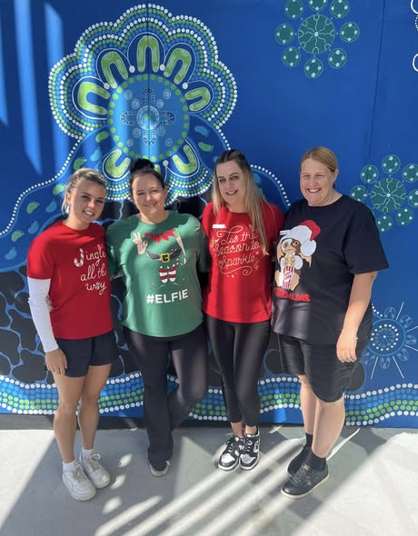 Tillys Cooranbong Four people stand smiling against a vibrant blue and green patterned wall. They wear festive shirts and casual shorts, conveying a cheerful holiday spirit.