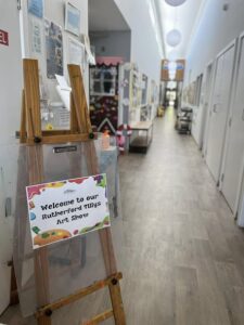 "A brightly lit hallway features a wooden easel with a colorful sign saying 'Welcome to our Rutherford Tillys Art Show.' The corridor has white walls, hardwood floors, and artwork displayed along the left wall, creating an inviting and creative atmosphere."