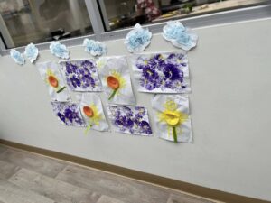 Art display with handmade flowers and clouds. Eight sheets feature purple dandelion prints and cheerful sunflowers, set against a white wall.