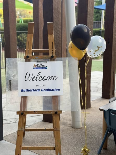 Tillys Rutherford Wooden easel displays a "Welcome to our Rutherford Graduation" sign. Beside it, black, yellow, and white balloons add a celebratory touch.