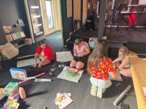 Tillys Waratah Four people sit on a carpeted floor, wrapping gifts in colorful paper. The setting is cozy, with books on shelves and a festive atmosphere.