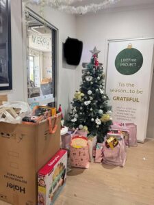 Tillys Waratah Christmas tree adorned with white and gold ornaments stands in a room filled with gift bags and boxes. Banner reads "LIVEFree PROJECT. Tis the season... to be GRATEFUL."