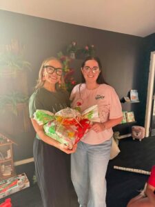 Tillys Waratah Two smiling women stand indoors holding a wrapped gift. One wears a green shirt, the other a pink shirt. The room has dark walls and festive decor.