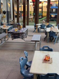 Tillys Rutherford Outdoor preschool area with small tables and chairs, decorated with lanterns and jars. The space feels festive and welcoming, with a playground visible.