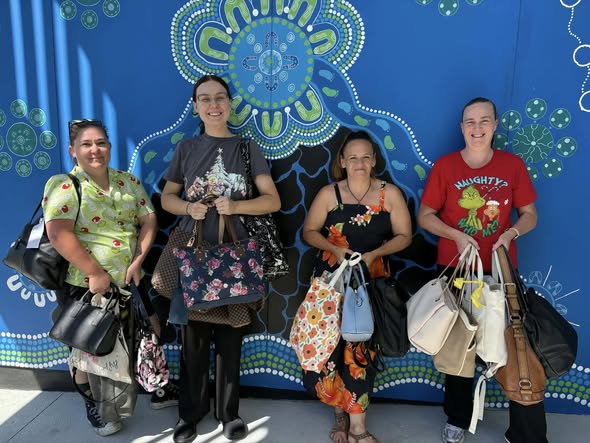 Four women smile and hold bags in front of a vibrant blue mural with circular patterns. They appear cheerful and proud, suggesting a community event.