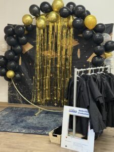 Tillys Rutherord Graduation-themed decoration with a backdrop of black and gold balloons, a fringe curtain, hanging graduation caps, and a rack of black gowns. Celebratory tone.