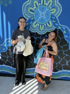 Tillys Cooranbong Two people smiling, holding colorful bags in front of a vibrant blue mural with organic patterns. The mood is cheerful and welcoming.