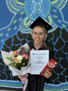 A young lady stands in front of a blue Aboriginal painted wall, wearing a black graduation gown and cap. She is holding a certificate, a kit-kat chocolate block and a colourful bunch of flowers. The image is bright and warm.