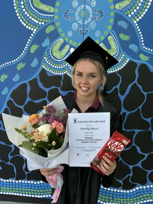 A young lady stands in front of a blue Aboriginal painted wall, wearing a black graduation gown and cap. She is holding a certificate, a kit-kat chocolate block and a colourful bunch of flowers. The image is bright and warm.
