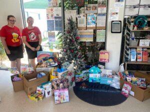Tillys Cardiff Two people in festive shirts stand beside a decorated Christmas tree indoors. Surrounding them are numerous colorful gift boxes and bags, creating a cheerful, holiday atmosphere.