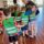 A group of children in matching green vests huddle around a smiling adult, conveying warmth and joy in a bright room with wooden floors.