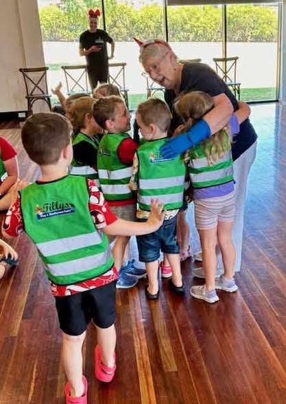 A group of children in matching green vests huddle around a smiling adult, conveying warmth and joy in a bright room with wooden floors.