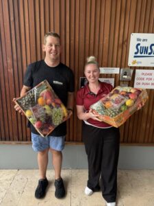Two people smile, holding fruit baskets, standing in front of a wooden wall. Both wear casual attire, and the mood is cheerful and celebratory.