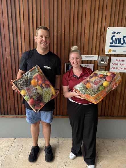 Two people smile, holding fruit baskets, standing in front of a wooden wall. Both wear casual attire, and the mood is cheerful and celebratory.