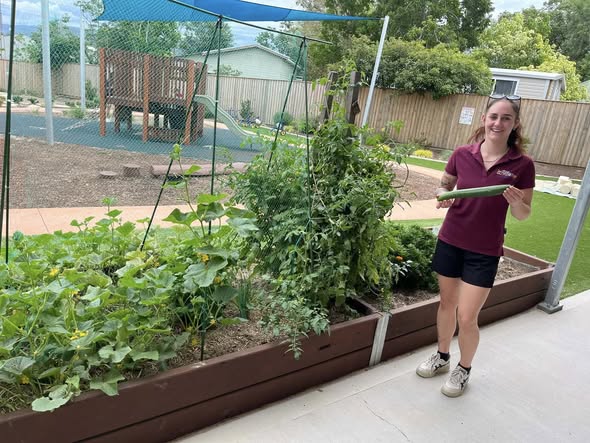 A smiling woman in a burgundy shirt holds a large cucumber, standing beside a lush vegetable garden in a sunny backyard with a play area.