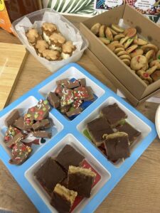 A table holds a variety of treats: gingerbread men, sprinkle-topped chocolate pieces, M&M cookies, and layered chocolate-peanut butter bars on colorful plates.