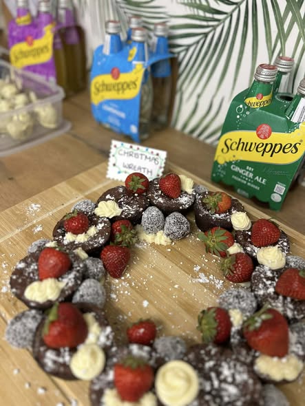 A festive chocolate wreath adorned with strawberries and cream sits on a wooden board. Nearby are Schweppes drink bottles and a sign reading "Christmas Wreath."