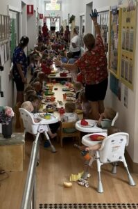 A bustling daycare hallway with toddlers seated at long tables, enjoying a colorful meal. Caregivers assist, creating a lively and joyful atmosphere.
