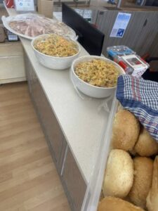 Alt text: "Bowls of pasta salad and a tray of fresh rolls on a countertop, with cured meats in the background. Bright and inviting indoor setting."