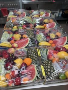 A table displaying multiple fruit trays wrapped in plastic. Each tray contains colorful fruits like watermelon, pineapple, bananas, and grapes, in a kitchen setting.