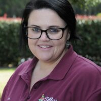 A young lady with dark brown hair and glasses smiles for a portrait. She is wearning a maroon work shirt, with a 'Tillys' logo on the front. There are trees and greenery in the background. The image it outside in a bright, summery setting.
