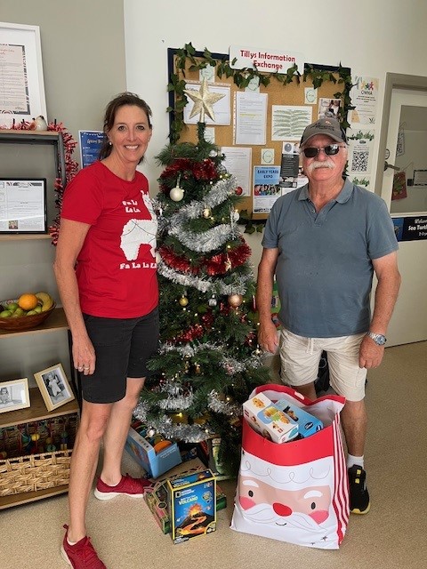 Two people stand in a foyer alongside a decorated Christmas Tree and gifts on the floor. There are posters and information sheets on the wall behind. The tone is cheerful and merry.