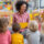A smiling preschool teacher sits on the floor with a small group of children. The background is colourful set within a classroom.