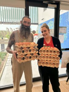 A smiling man and woman hold large stacks of egg cartons in a bright room. The background features window decals and a mural, evoking a cheerful tone.