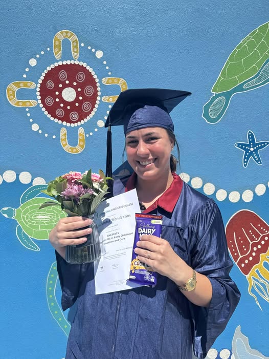 Tillys Waratah Graduate in blue cap and gown holds flowers, a certificate, and chocolate, smiling against a colorful mural of sea creatures. Celebratory and joyful tone.